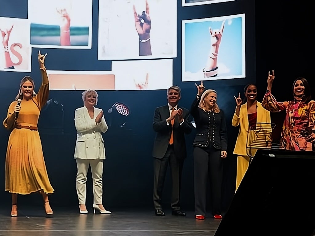 Kendra Scott and five others hold up the Hook 'Em Horns hand sign on stage at an event.